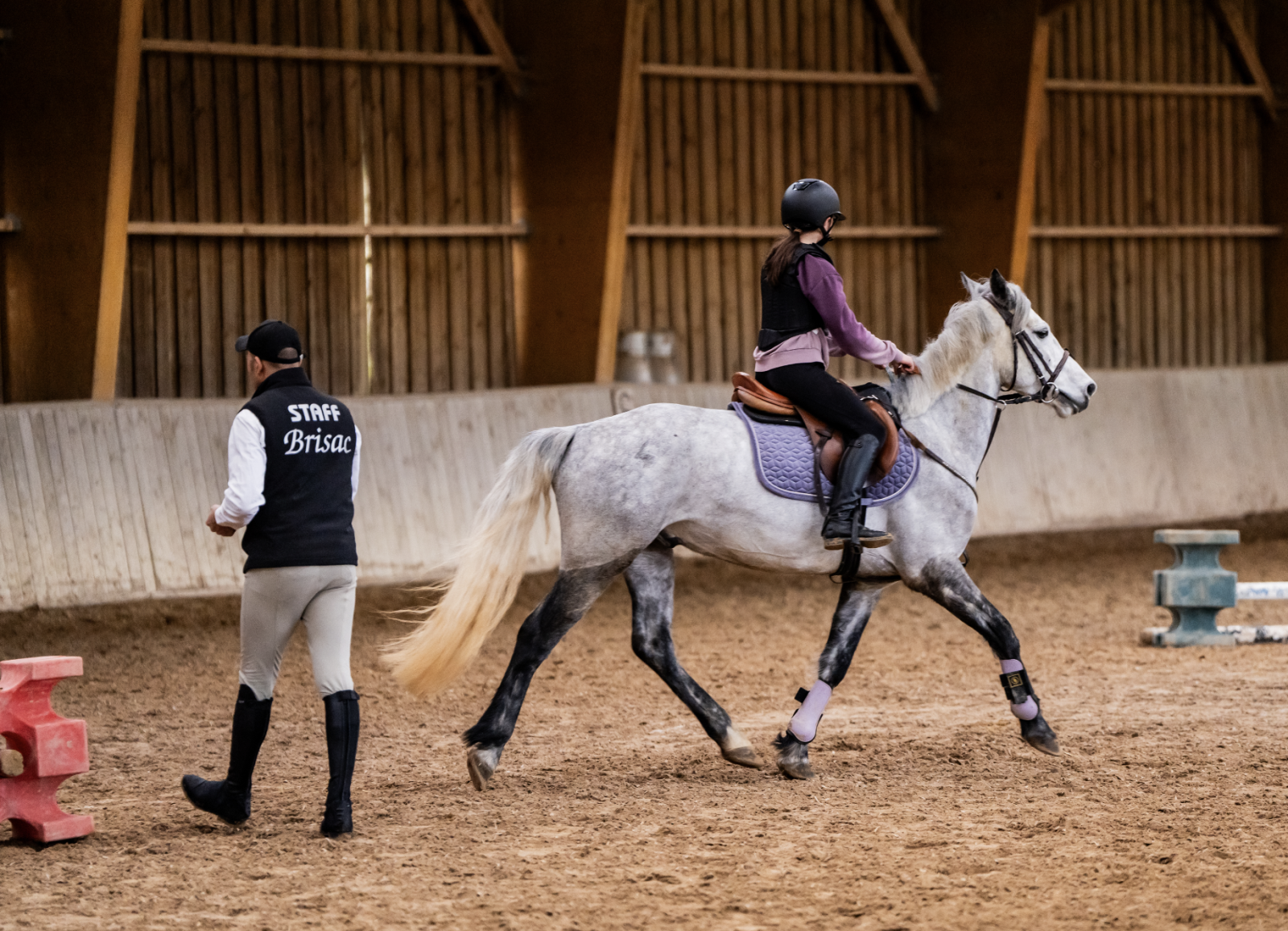 Une monitrice donne des indications à une cavalière qui monte un cheval gris. Brisac Equiteam